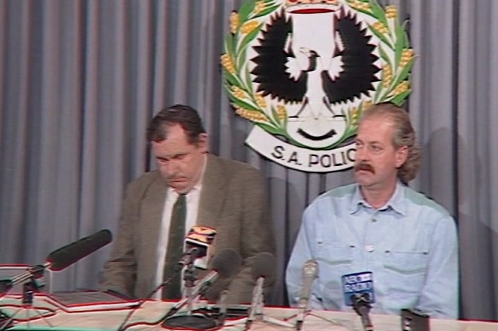 A police officer wearing a suit sitting next to Leon Barreau behind a table with microphones sitting on it