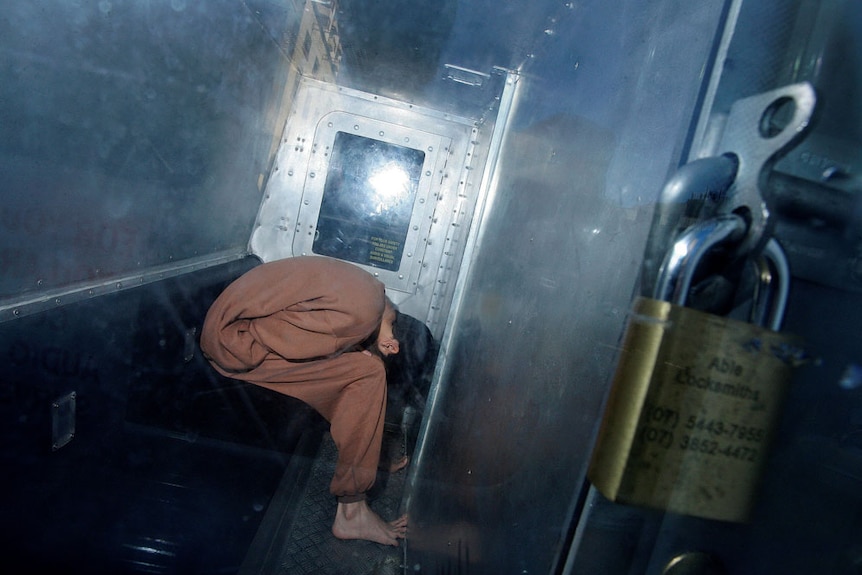 A man in long brown gown is doubled over in a padlocked metal police prison van, light reflects off the glass door.