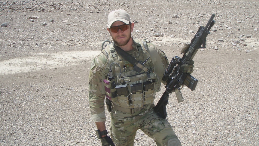 A soldier in uniform and sunglasses holds his rifle in a dry and dusty environment.