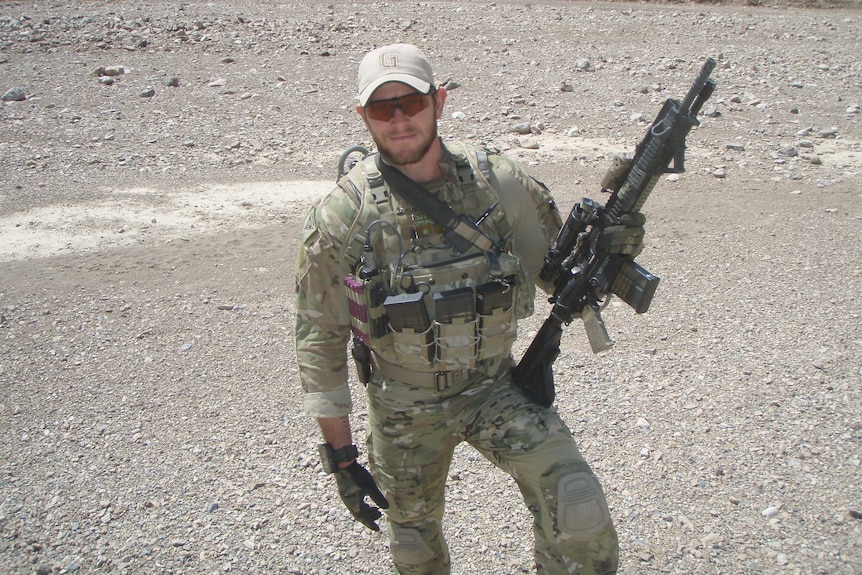 A soldier in uniform and sunglasses holds his rifle in a dry and dusty environment.