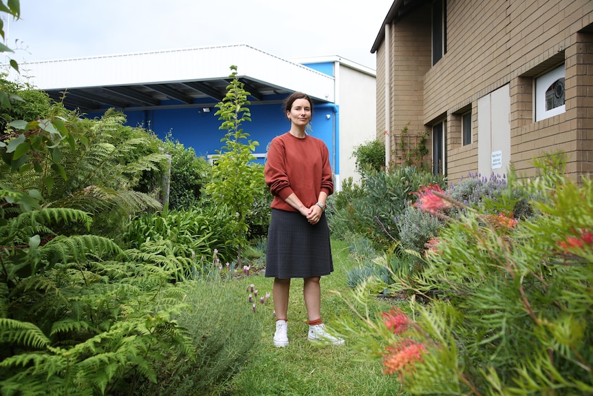 Lara standing in her garden behind her house, among thriving and flowering plants.