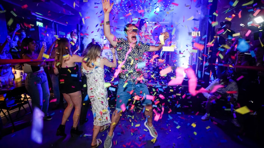 A man wearing a colourful shirt and shorts jumps in the air as pink streamers fall from the ceiling of a club.