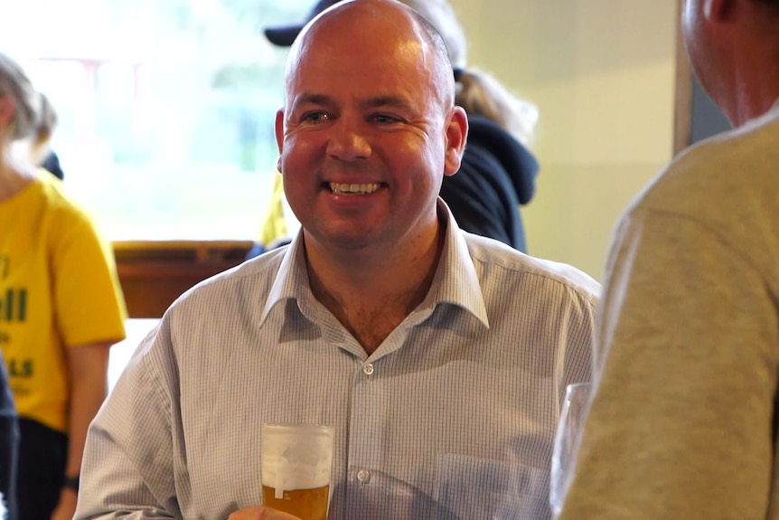 A bald man holding a beer smiles at someone off camera.