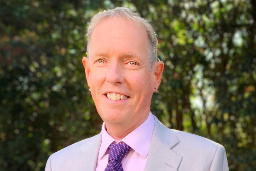 A middle-aged man in a grey suit, pink shirt and purple tie