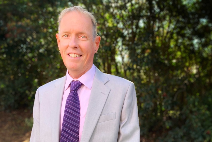 A middle-aged man in a grey suit, pink shirt and purple tie