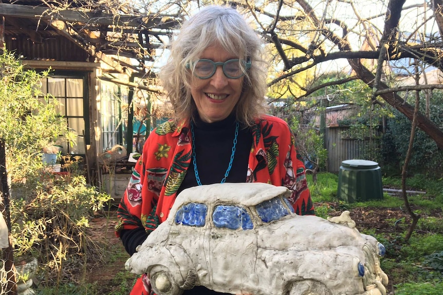 Elderly woman in glasses and a red patterned shirt holding a sculpture of a Holden and smiling