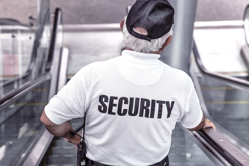 A man in a white t-shirt with the word "security" on the back rides down an escalator.