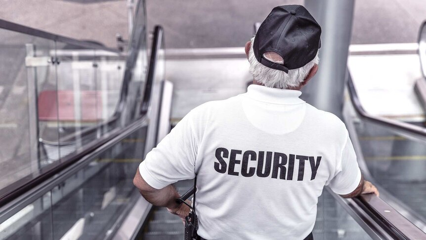 A man in a white t-shirt with the word "security" on the back rides down an escalator.