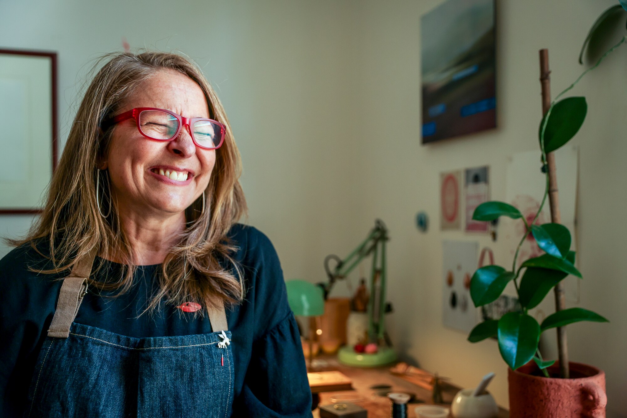 A woman in red glasses sits in a studio and screws her face up in laughter.