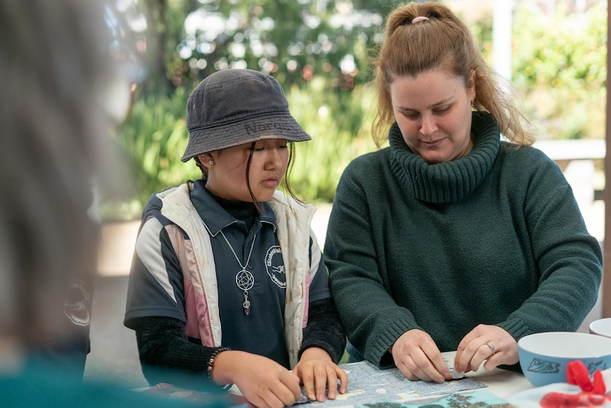 A child wearing a green school uniform and hat stands next to a woman wearing a green jumper, looking down at a table with craft