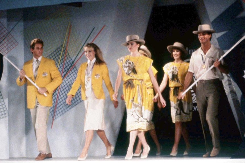 Four people walking onto an opening ceremony stage in yellow animal dresses and holding aussie flags