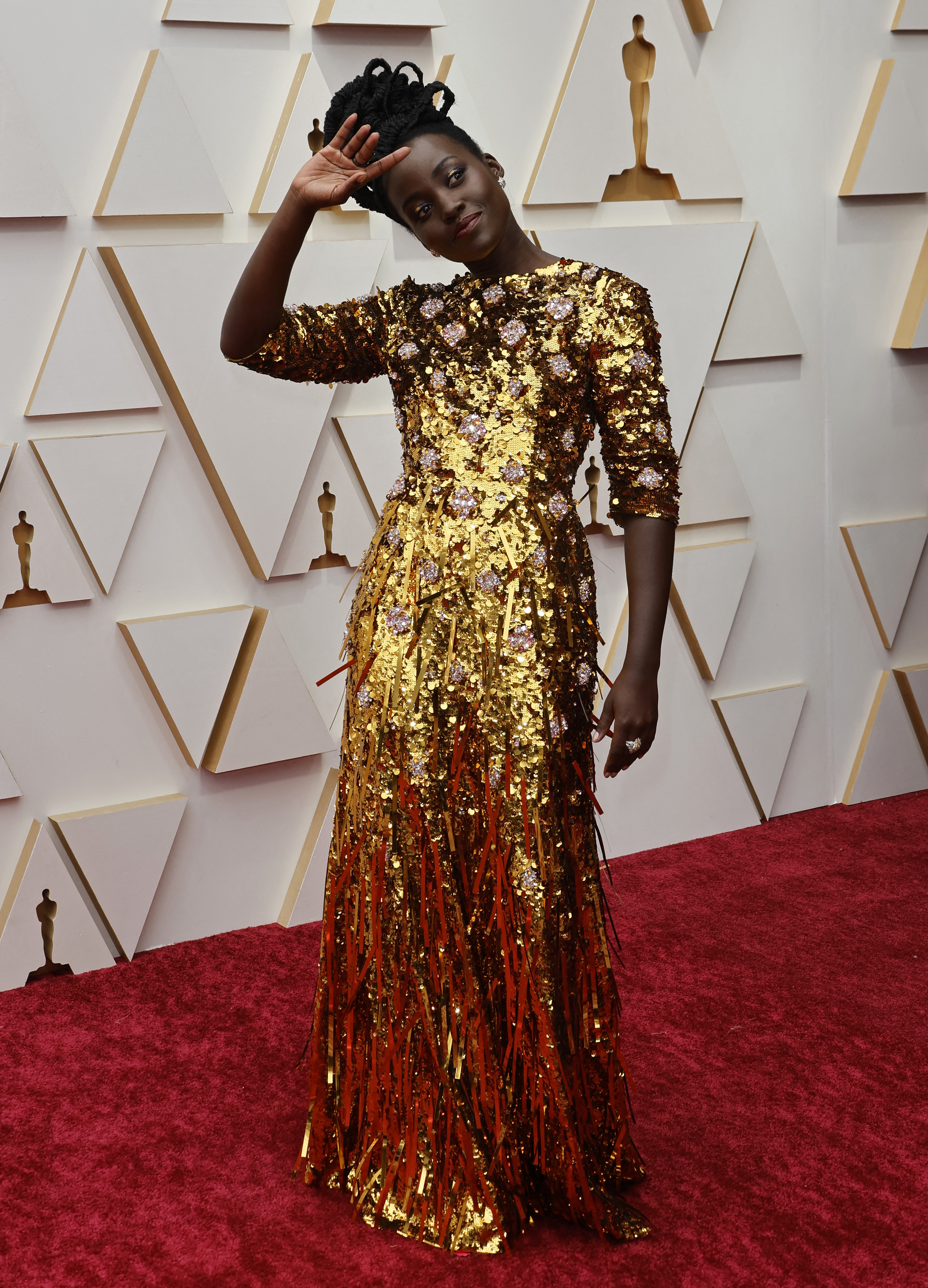 lupita nyong'o poses with her hand to her head on the oscars red carpet wearing a metallic-gold tasselled gown