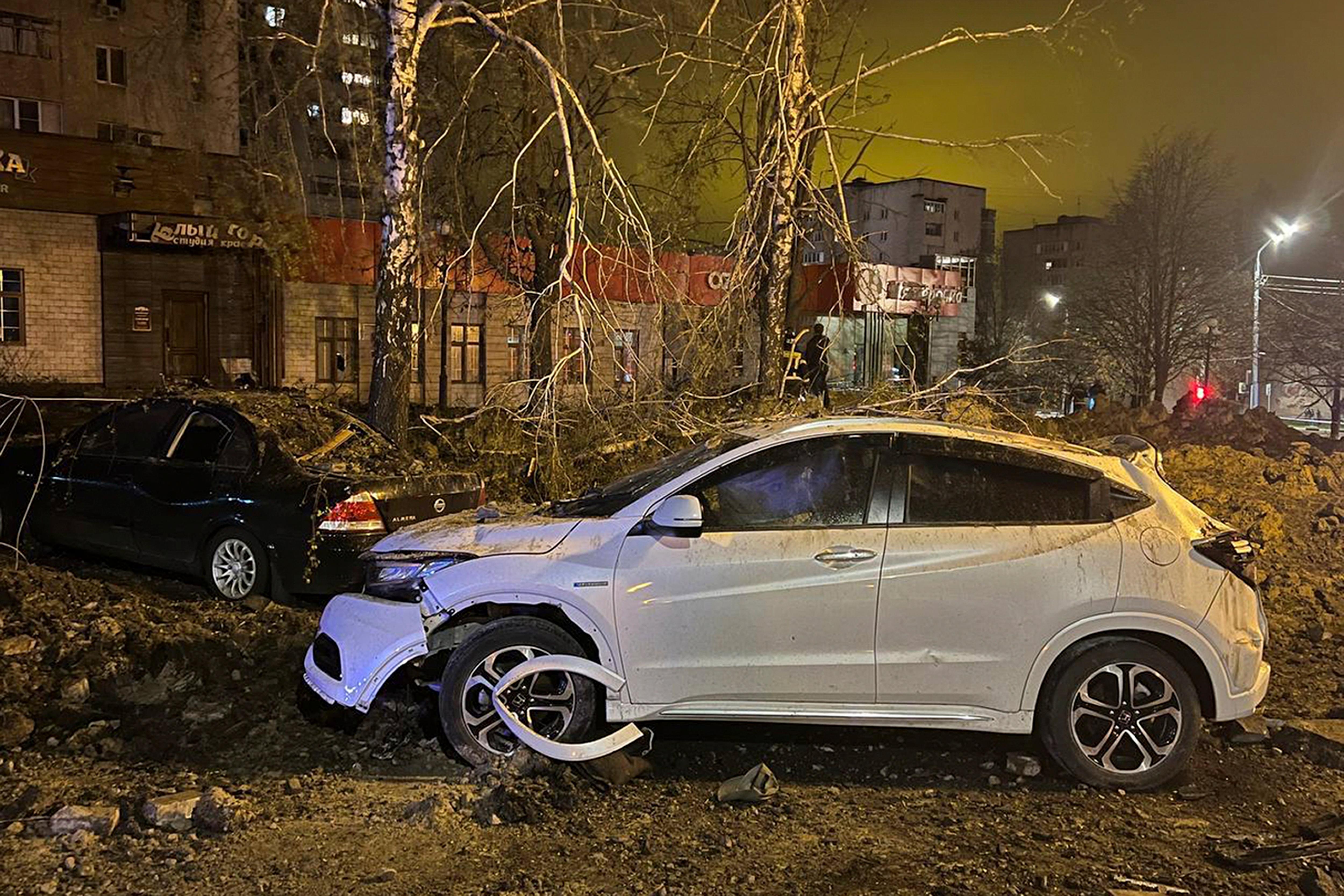 A white hatchback with damage front tires, roof and bonet sits near a crater created by a large explosion.