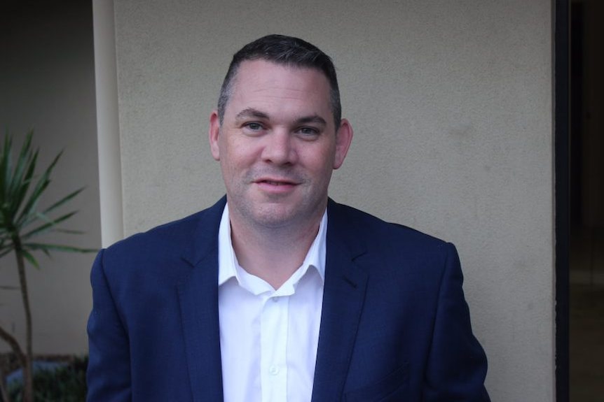 A middle aged white man wearing a white shirt and a suit jacket poses for a photo.