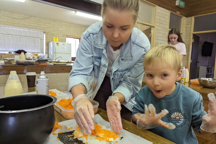 A blonde woman wearing a denim jacket helps an excited blonde boy make sushi rolls.