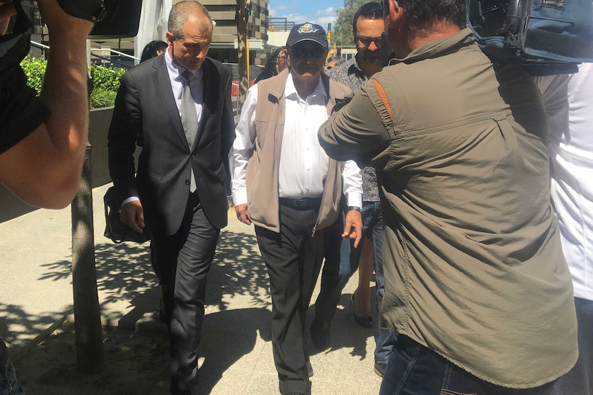 Keith Osman walks outside the Perth District Court flanked by a man in a suit with TV cameramen in front of him.