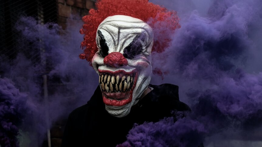 Picture of a scary clown with curly red hair smiling with big sharp teeth in front of purple smoke