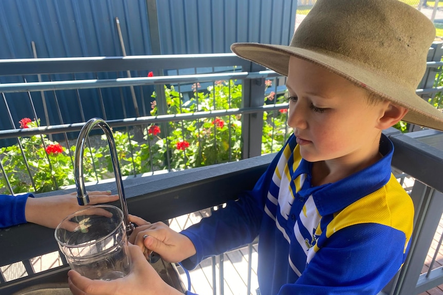 A primary school boy in a blue uniform standing at a tap, filling a glass.