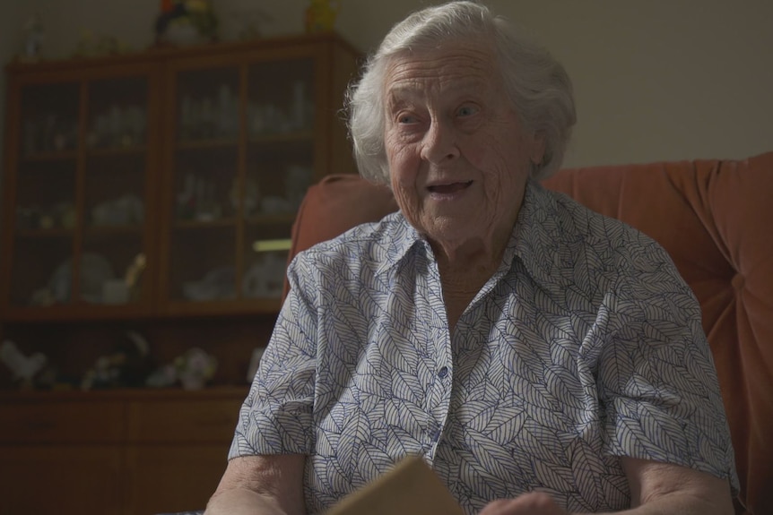 An elderly woman win a blue dress sits on a sofa with a sideboard behind her