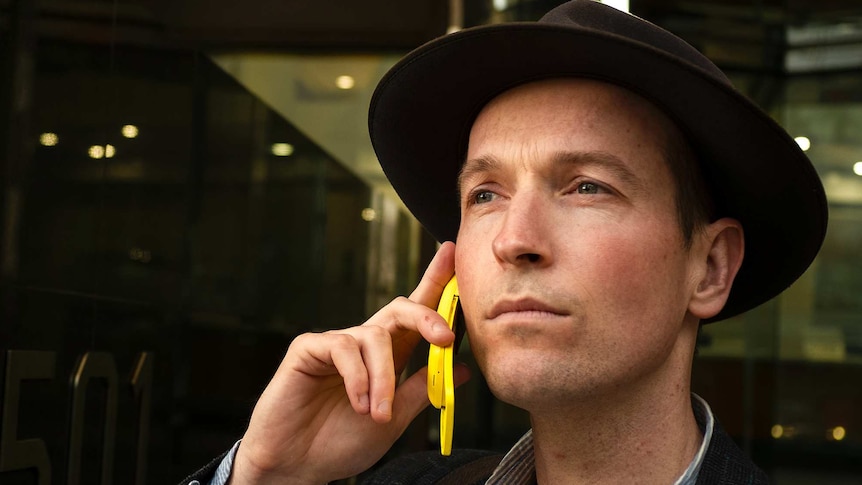 Jesse Noakes standing in a street wearing a hat and using his yellow Nokia flip phone.