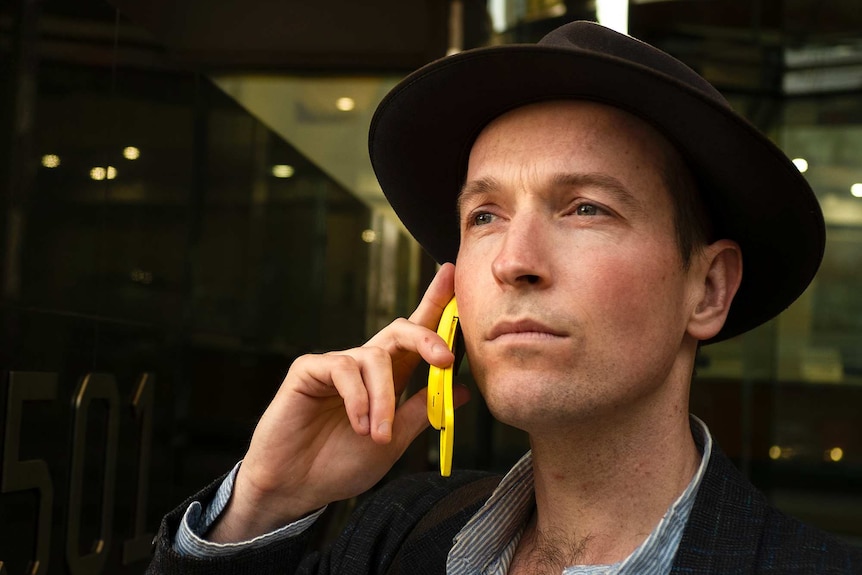Jesse Noakes standing in a street wearing a hat and using his yellow Nokia flip phone.