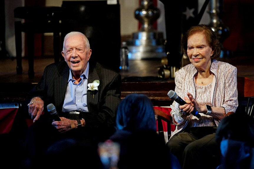 A medium shot of an elderly man and elderly woman sitting near each other indoors, both holding microphones and smiling