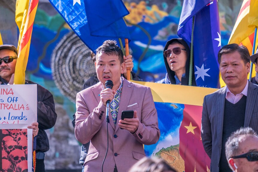 Looking up at a protest crowd, you see a man in a salmon-coloured blazer speaking into a microphone and holding a smartphone.