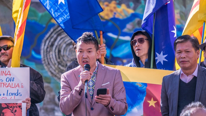 Looking up at a protest crowd, you see a man in a salmon-coloured blazer speaking into a microphone and holding a smartphone.