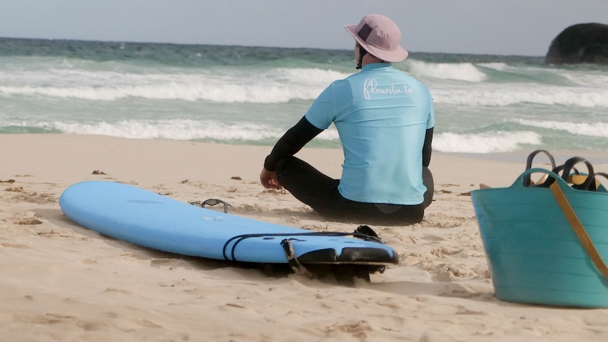 A man sitting on the sand looking out at the ocean with a surfboard next to him 