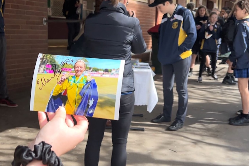 A student holds a signed photo of Ellen Ryan at the Commonwealth Games with students in the background.