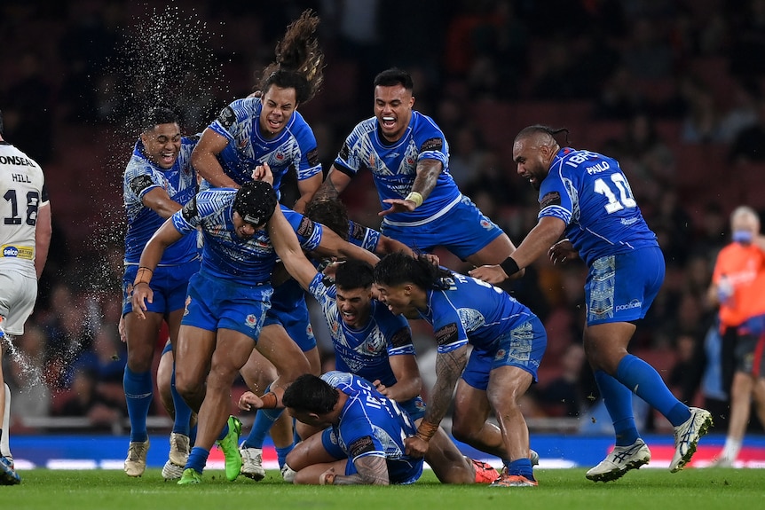 Sekelompok pemain liga rugbi Samoa berteriak dan menyeringai saat mereka melompat ke atas rekan satu tim setelah meraih kemenangan besar.