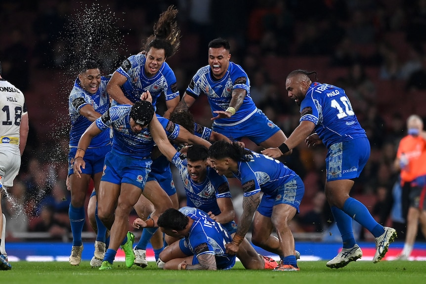 Un grupo de jugadores de la liga de rugby de Samoa gritan y sonríen mientras saltan sobre sus compañeros de equipo después de una gran victoria.