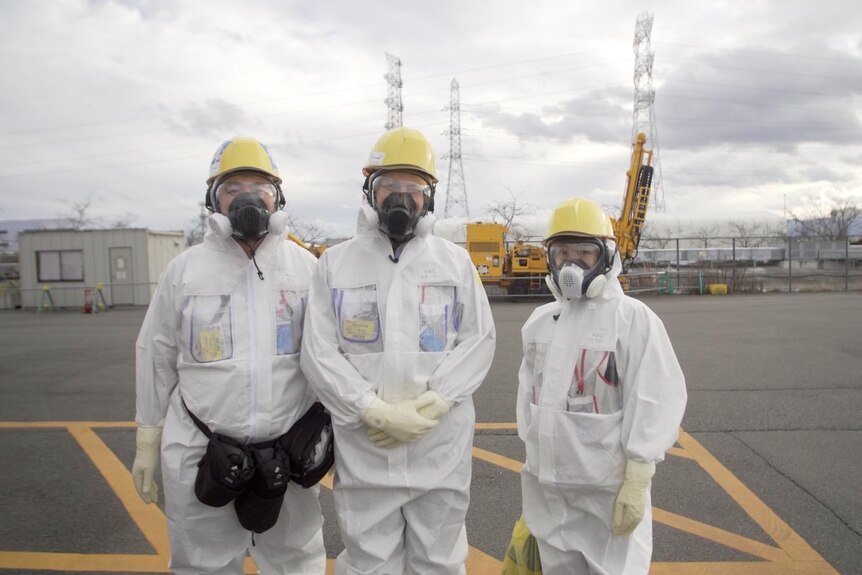 Three people standing in white suits, wearing breathing masks and helmets with facility in background.