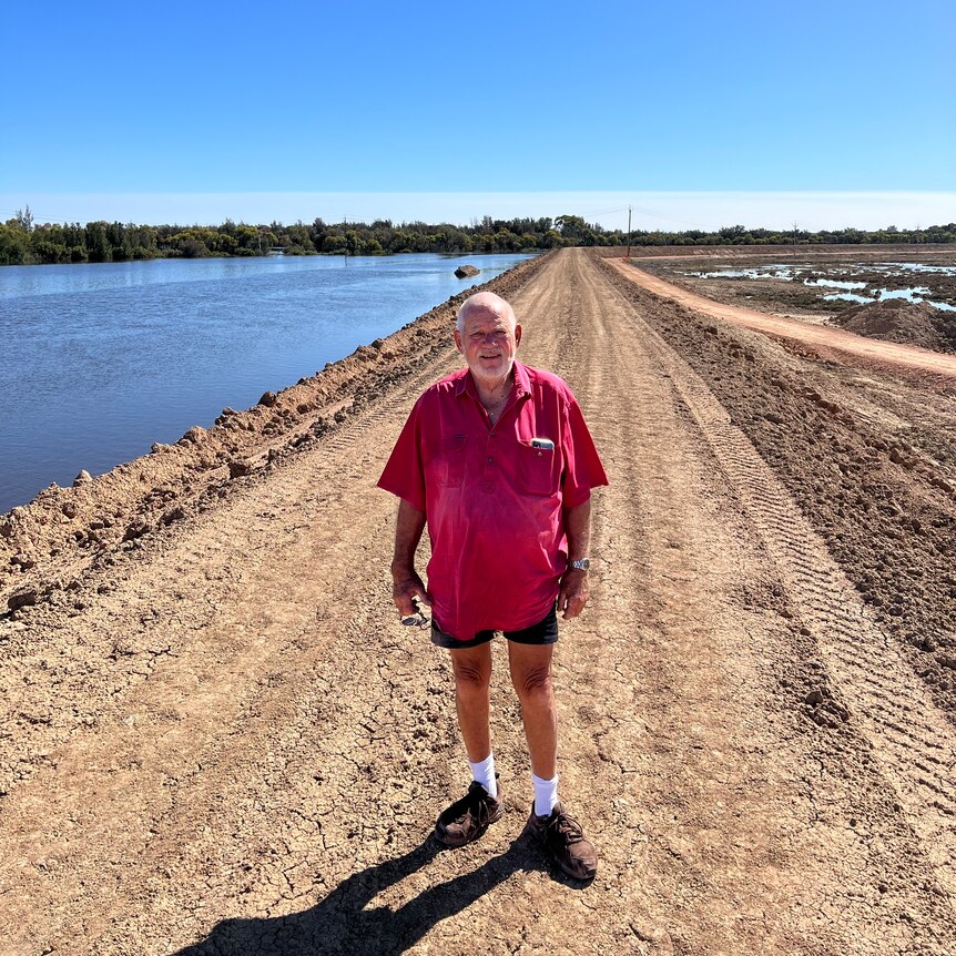 A man wearing a red shirt and shorts standing on a dirt levee next to a river