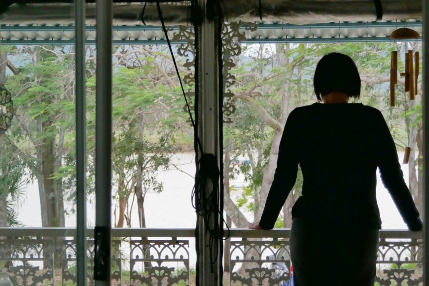 A woman stands on a front verandah and looks out towards a river