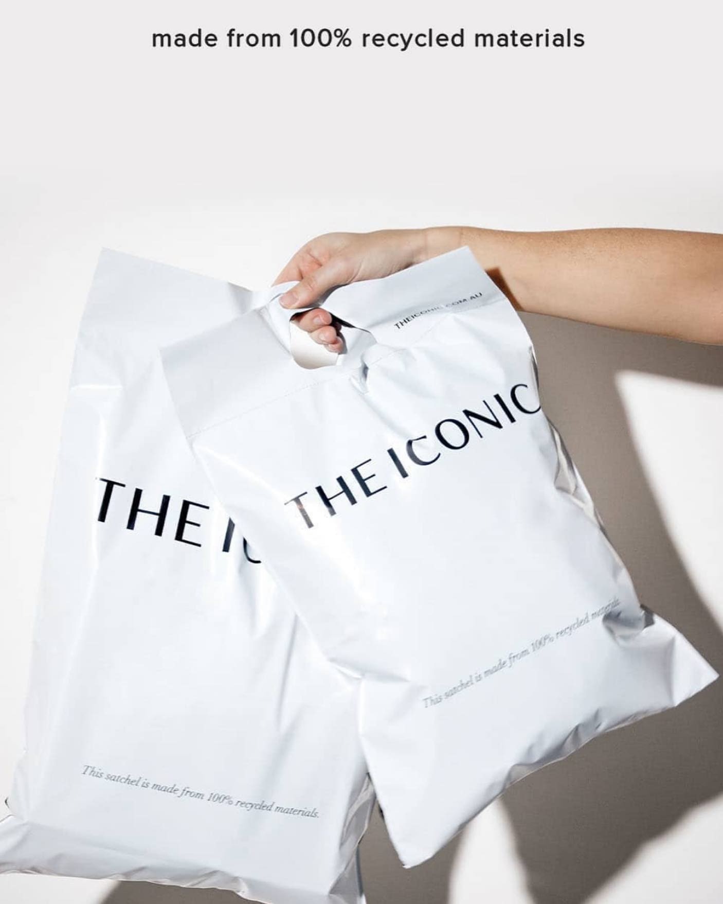 A woman's hand holds two large white satchels with "The Iconic" written on the front in black.