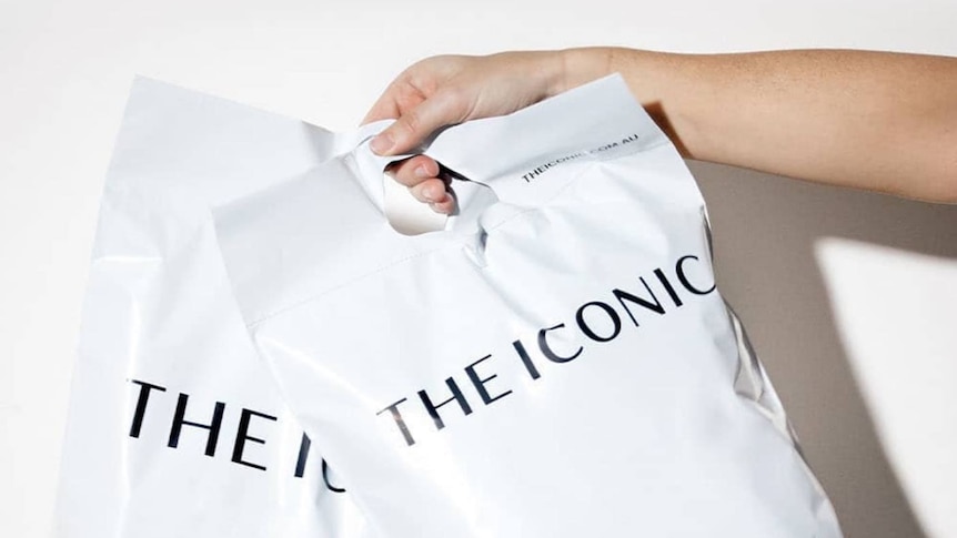 A woman's hand holds two large white satchels with "The Iconic" written on the front in black.