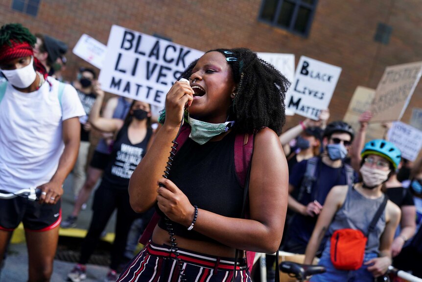 A young black woman talks into a hand-held microphone in a rally with a crowd of people behind.