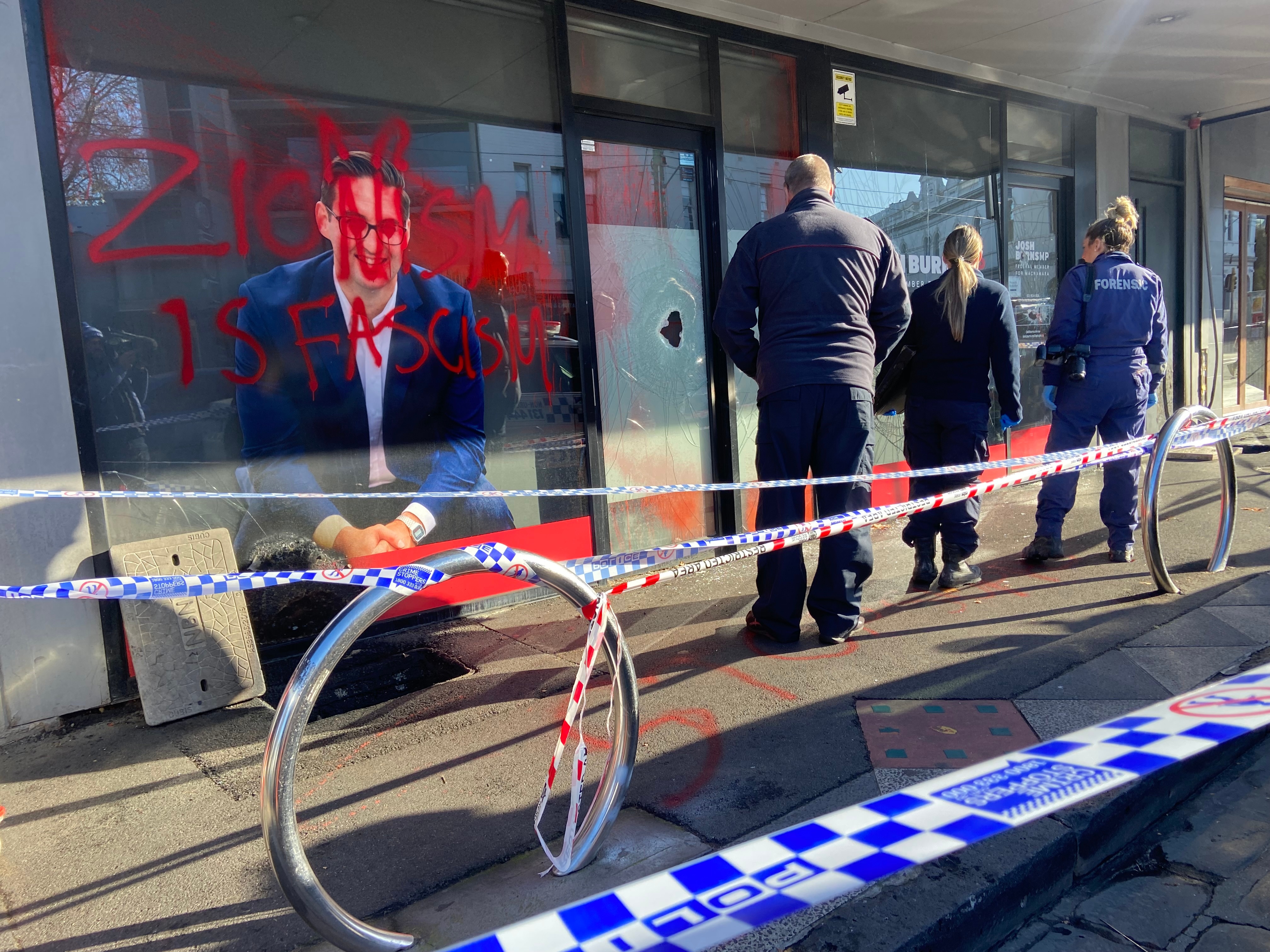 Police stand outside an office, covered in red paint reading 'ZIONISM IS FASCISM', with smashed windows.
