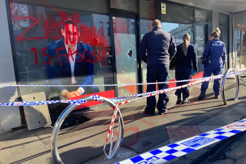Police stand outside an office, covered in red paint reading 'ZIONISM IS FASCISM', with smashed windows.