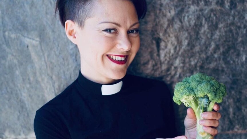 smiling young woman with cropped hair wearing clerical collar and black priest clothes holding a broccoli.