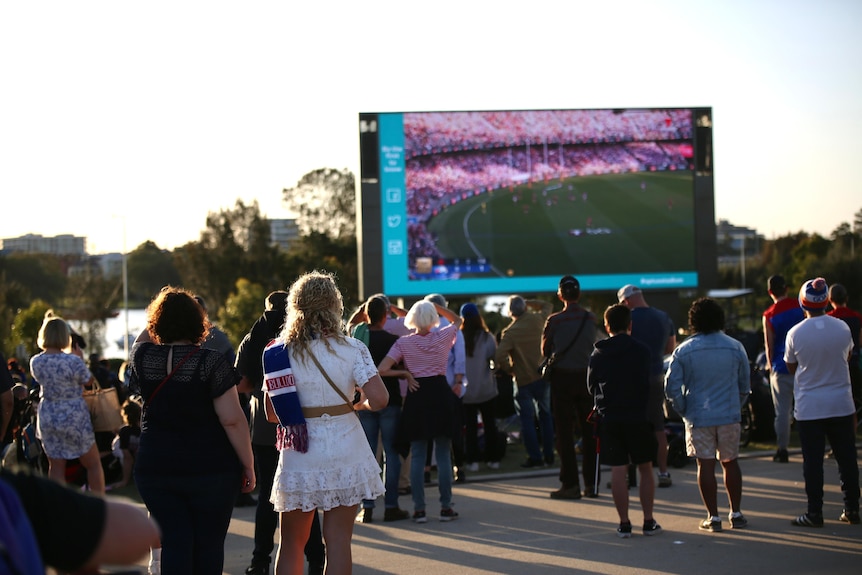 A crowd of people wearing Demons and Bulldogs attire stand before a large screen broadcasting a football match.