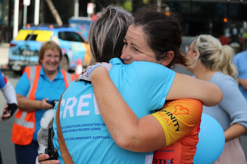 A woman in a cycling suit is emotional as she embraces a woman with the words "CREW" on the back of her shirt.