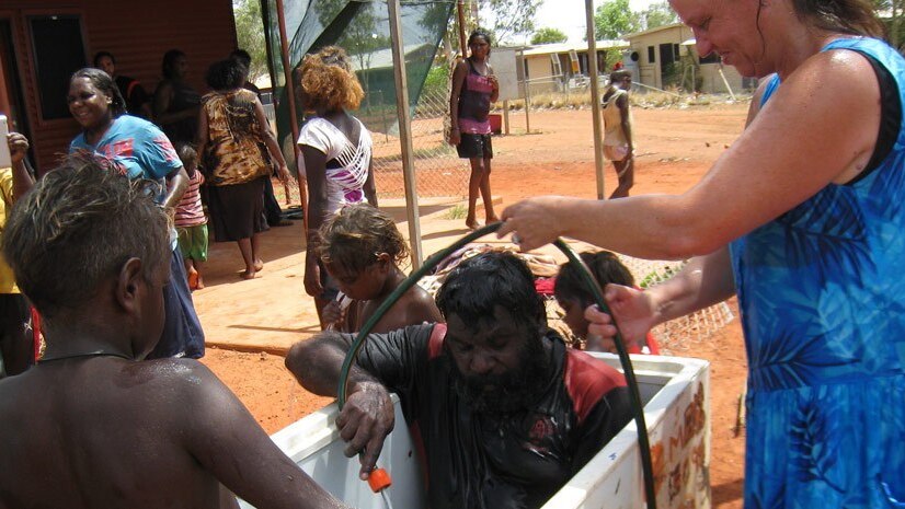An Aboriginal man sits in a bathtub, with a white woman standing above him handing him a hose. He is holding the end of the hose