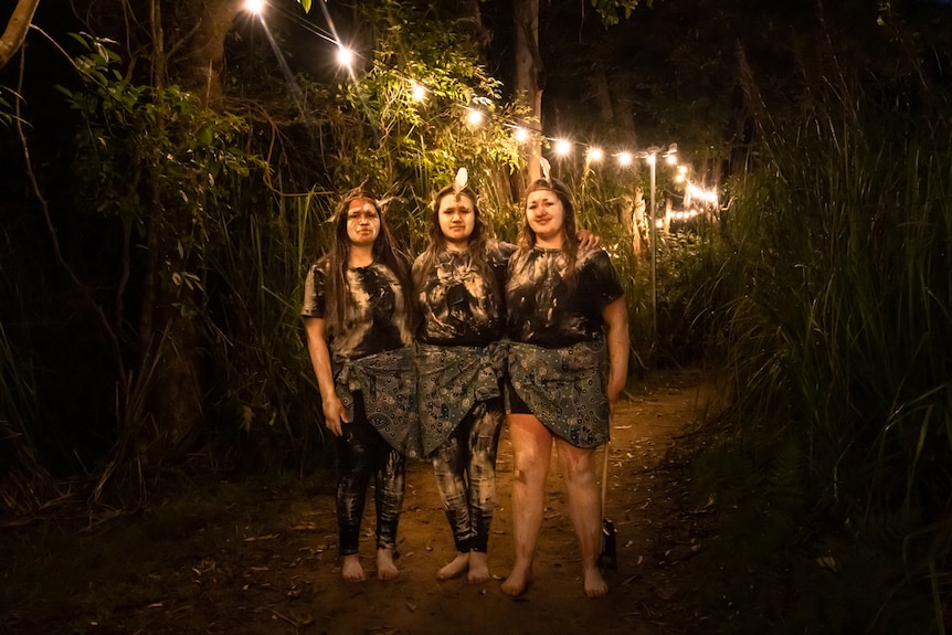 Three Indigenous teenagers in traditional dress and ochre standing on a lit path in a forest