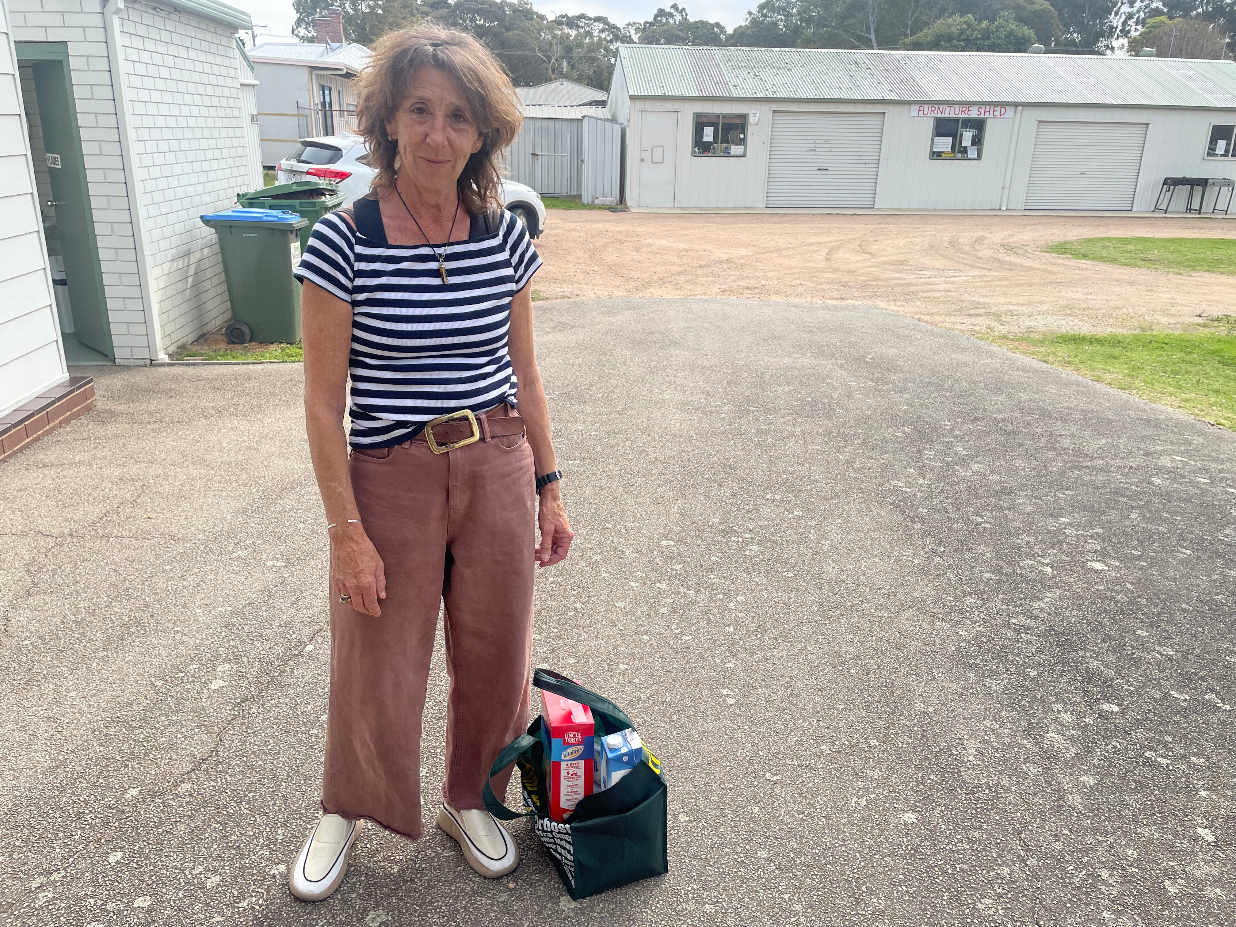 An older woman with shaggy dark hair cut shot, wears striped black and white tee, stands next to her bag of milk and cereal.