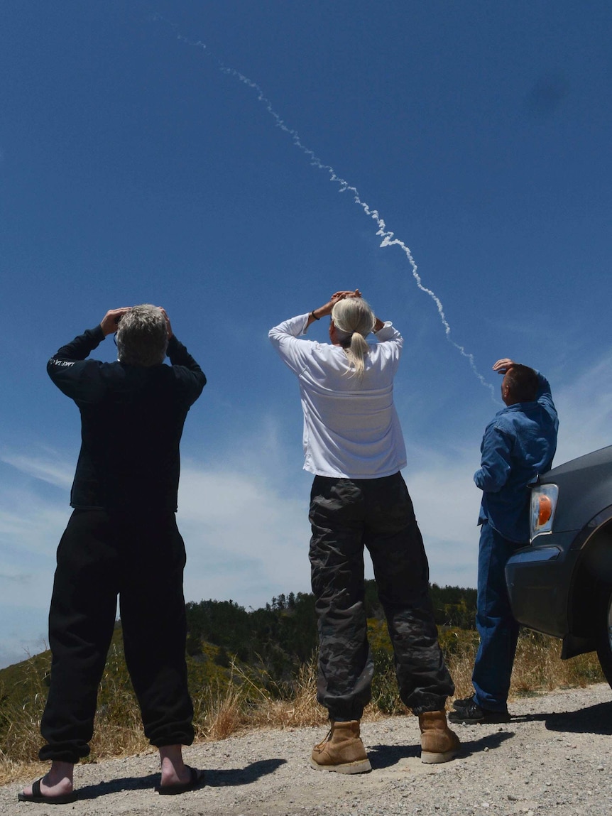 Three spectators watch an interceptor missile traverse a blue sky.