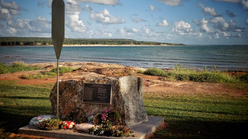 Memorial stone with plaque is surrounded by flowers and located overlooking a bay.