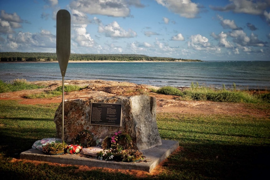 Memorial stone with plaque is surrounded by flowers and located overlooking a bay.