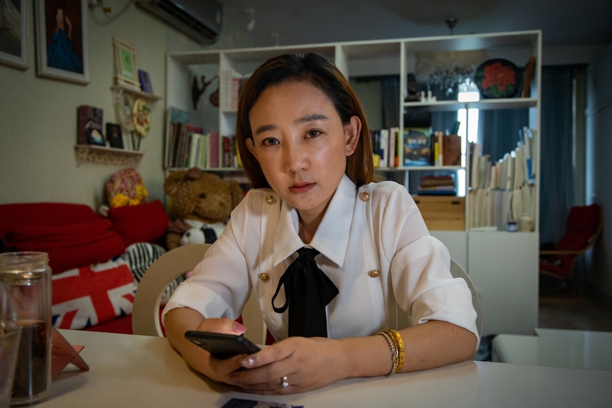 A young woman sits at a table in an apartment.